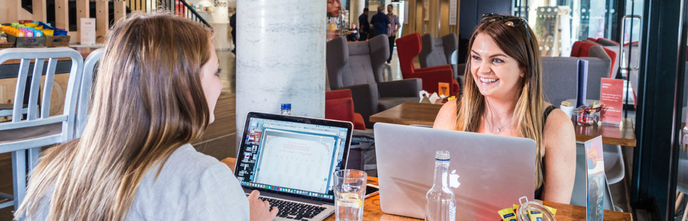 Two people sat working on laptops at a table talking and laughing